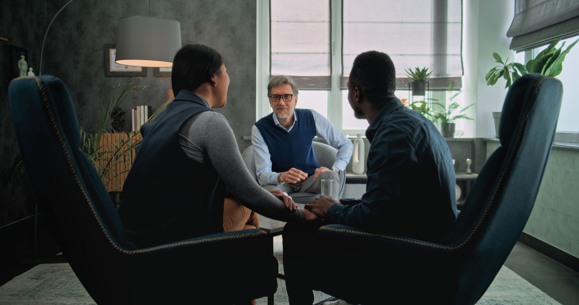 A man and a woman are sitting in chairs holding hands while talking to a counselor.