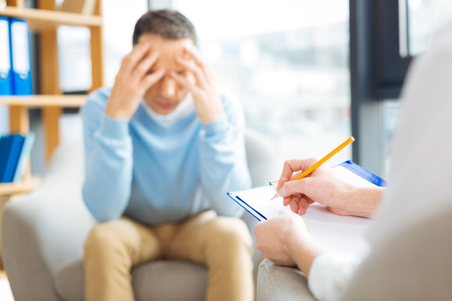 A man is sitting on a couch with his head in his hands while a woman writes on a clipboard.