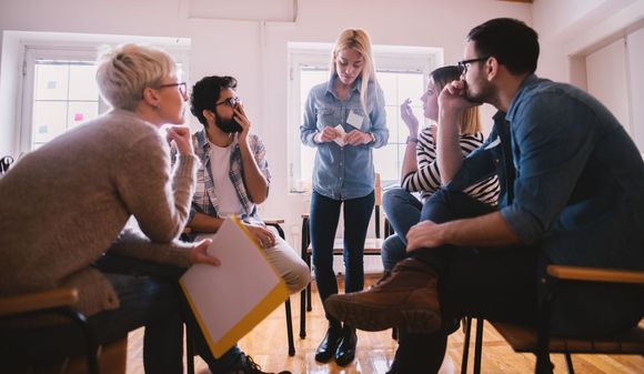 People in a circle, in a group meeting. A woman stands in the center talking; others listen. Bright room, wooden chairs.