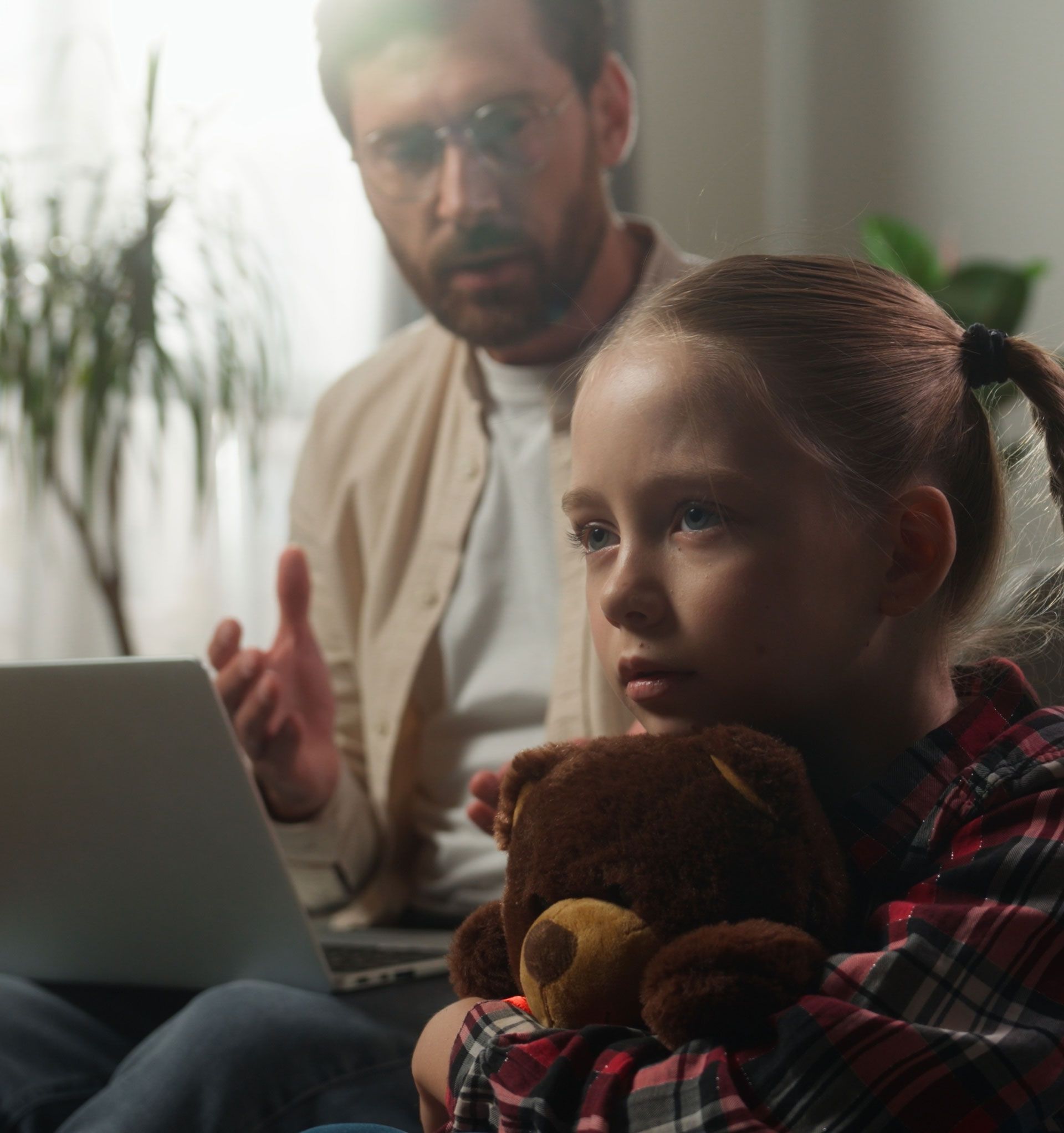 A little girl is holding a teddy bear while sitting in front of a laptop computer.