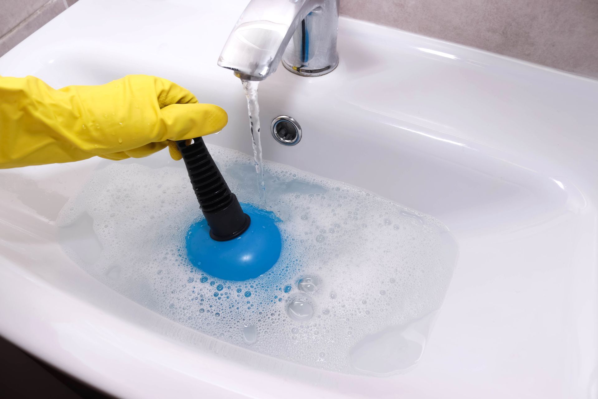 Yellow-gloved hand using a plunger in a foamy white sink under running water