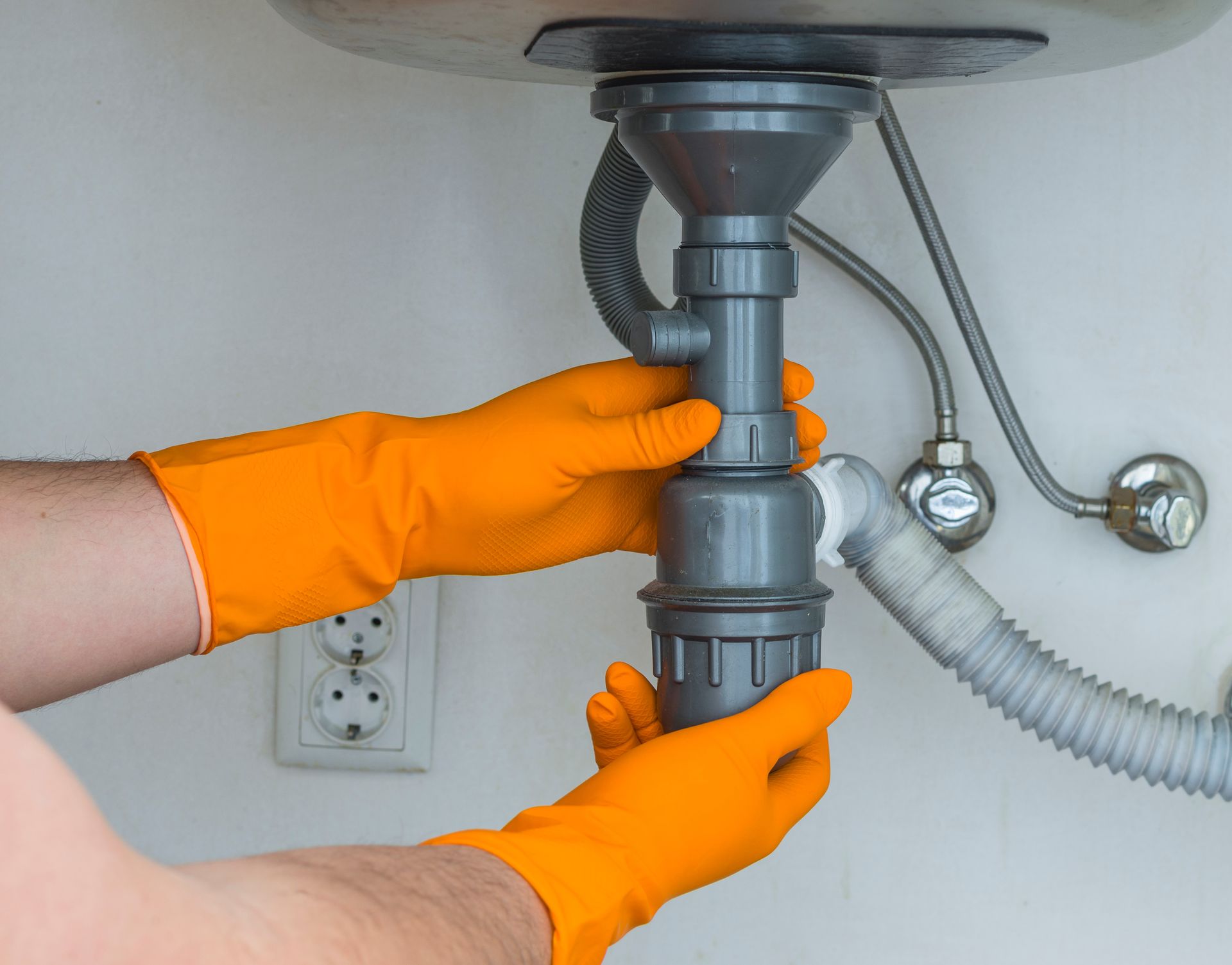 Orange-gloved hands tightening a gray sink drain pipe under a bathroom sink