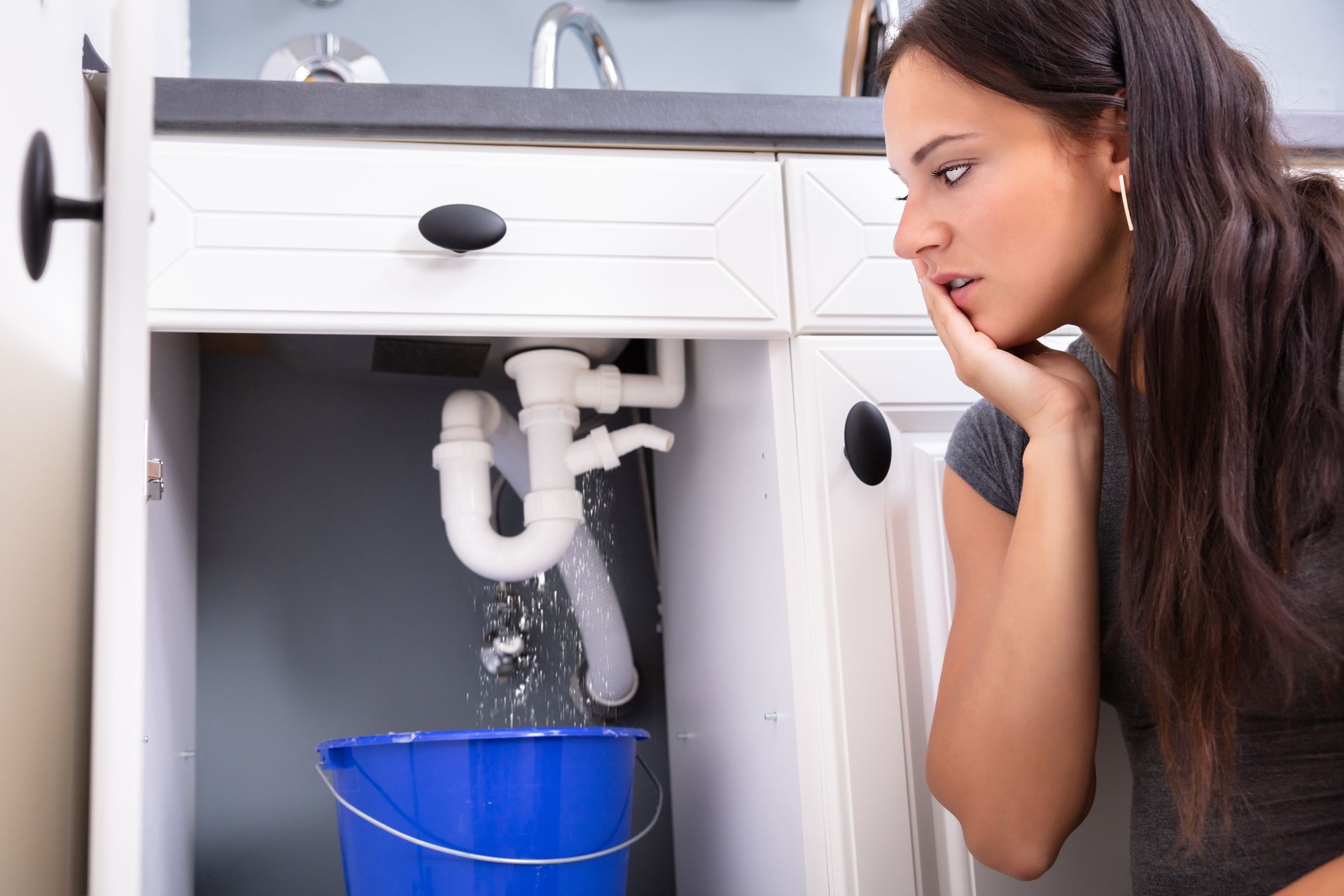 Woman looking under a sink at a leaking pipe into a blue bucket Woman looking under a sink at a leaking pipe into a blue bucket