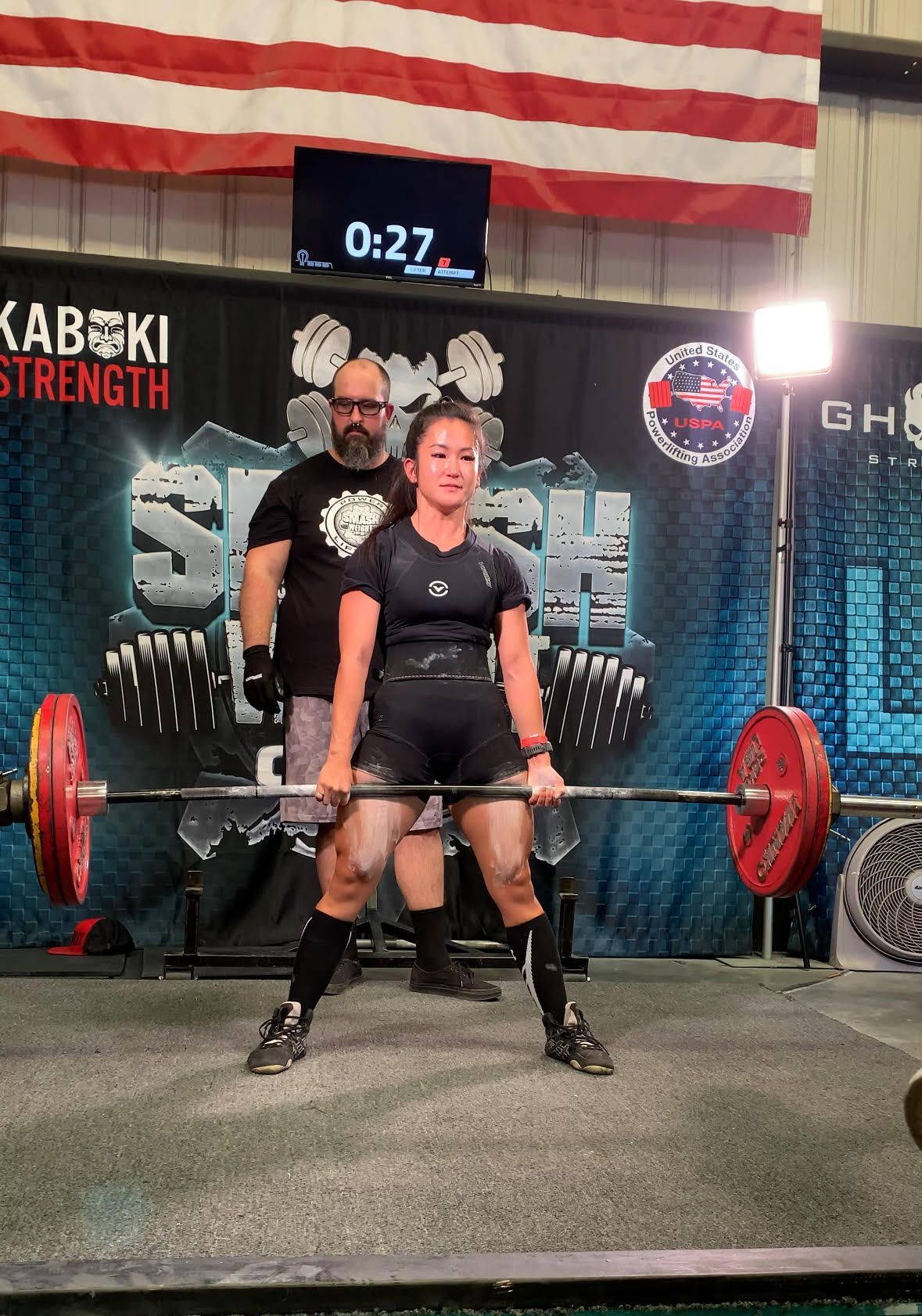 A woman is lifting a barbell in front of a kabuki strength sign