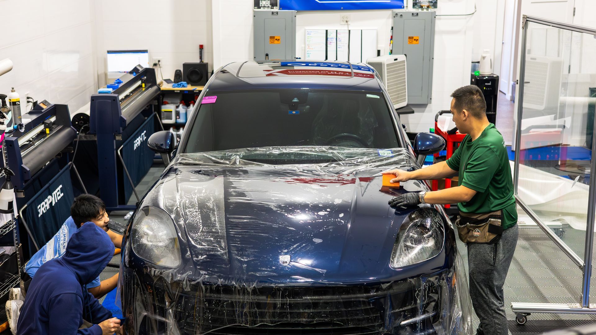 Workers applying protective wrap to a dark blue SUV in an auto shop