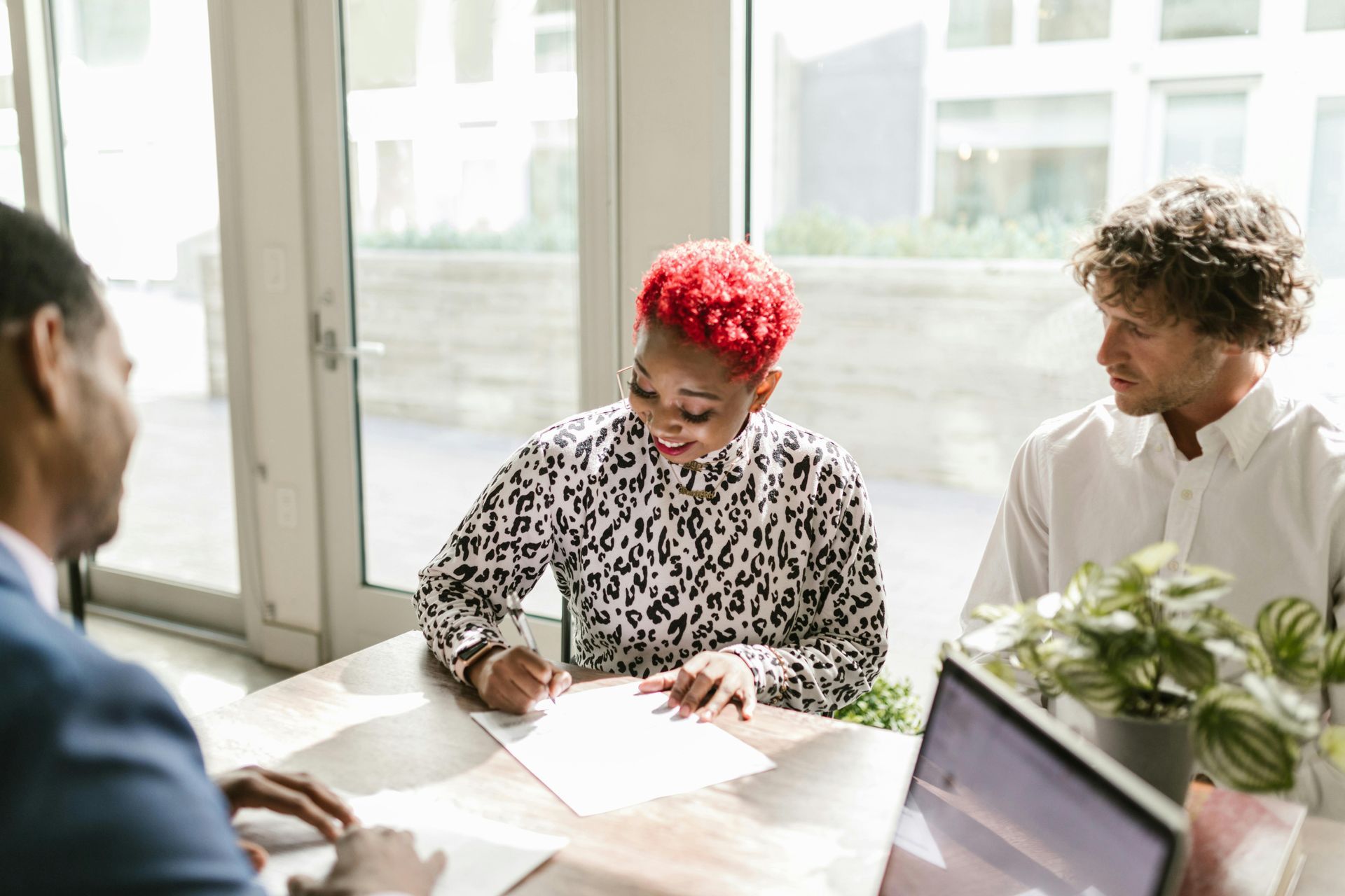 Three coworkers reviewing documents at a sunlit table with a laptop and plant nearby