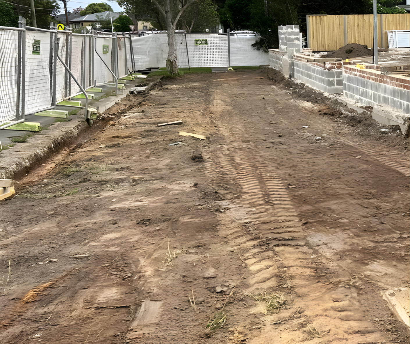 Bare dirt driveway or construction access path between white fences and stacked building materials.