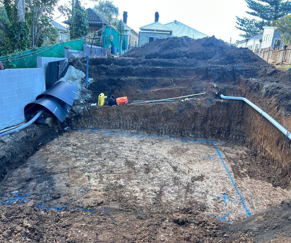 Excavated construction site with stepped dirt levels, blue foundation markings, and exposed pipes.