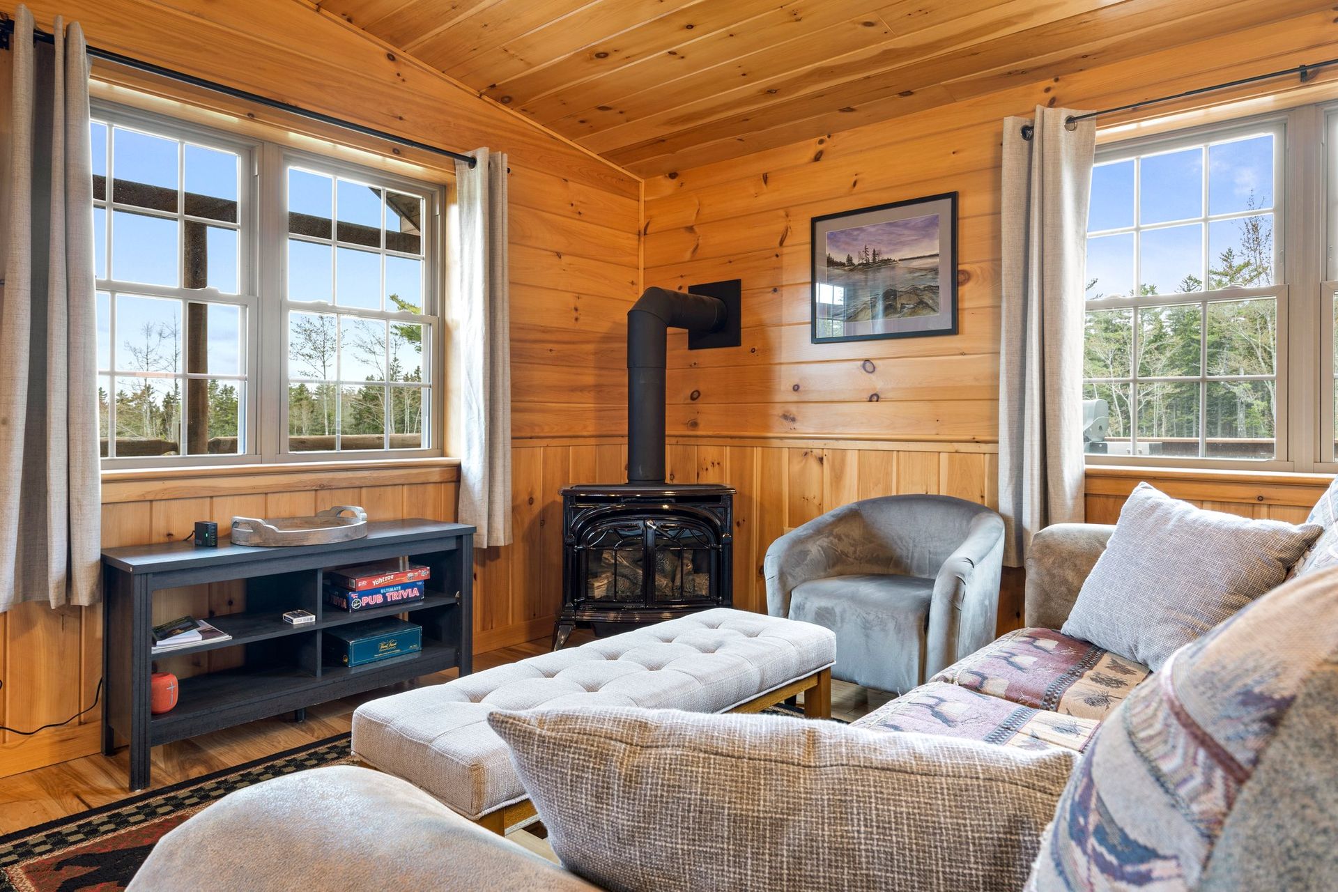 Cozy cabin living room with wood paneling, a stove, and a window view of trees. A couch and armchair offer seating.