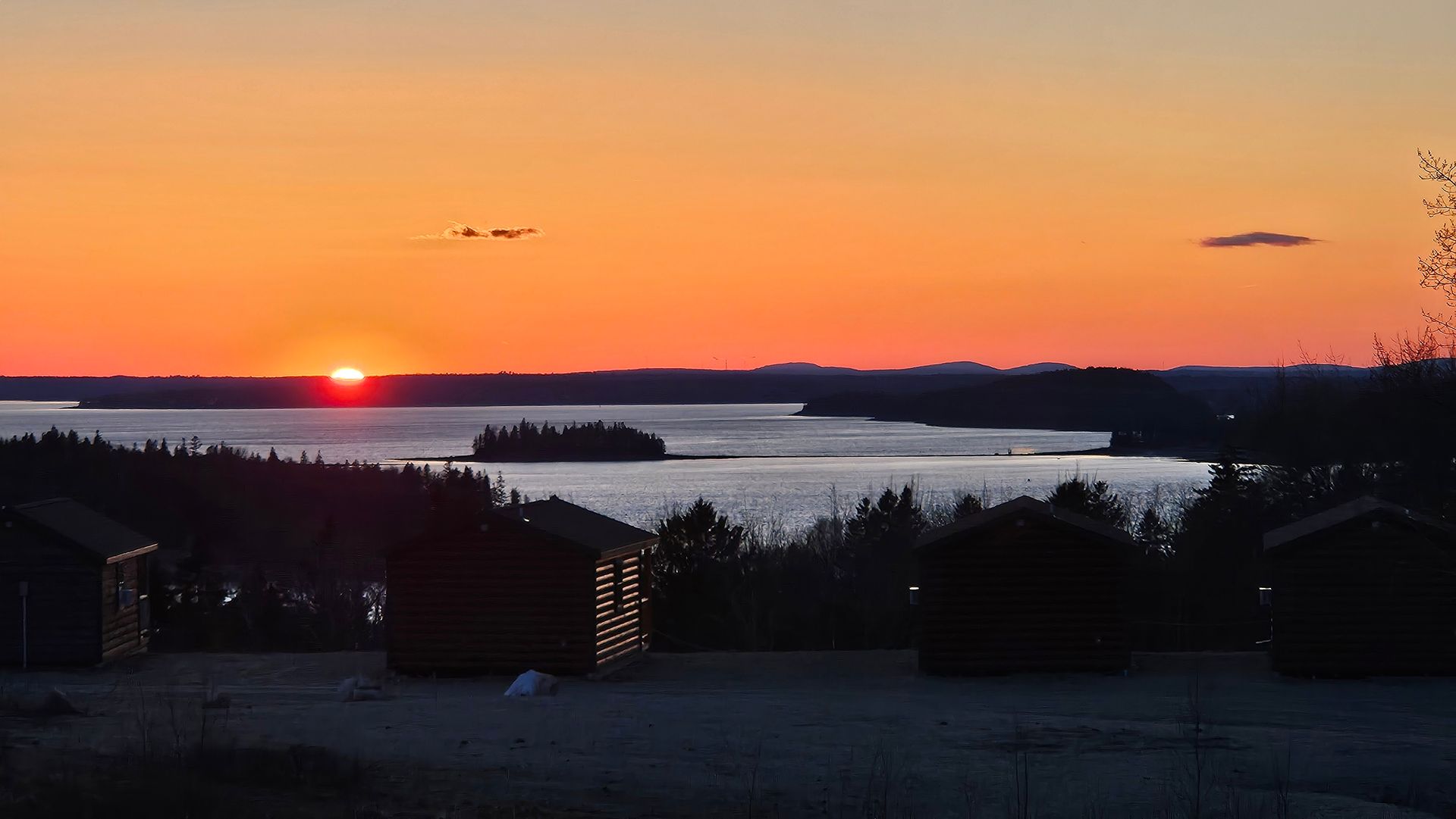 A sunset over a ocean with trees and cabins in the foreground