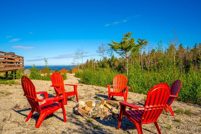 Red Adirondack chairs arranged around a fire pit on a gravel area, overlooking a forest and ocean under a blue sky.