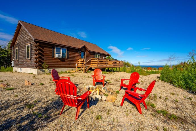 A log cabin with red Adirondack chairs around a campfire pit on a sunny day.