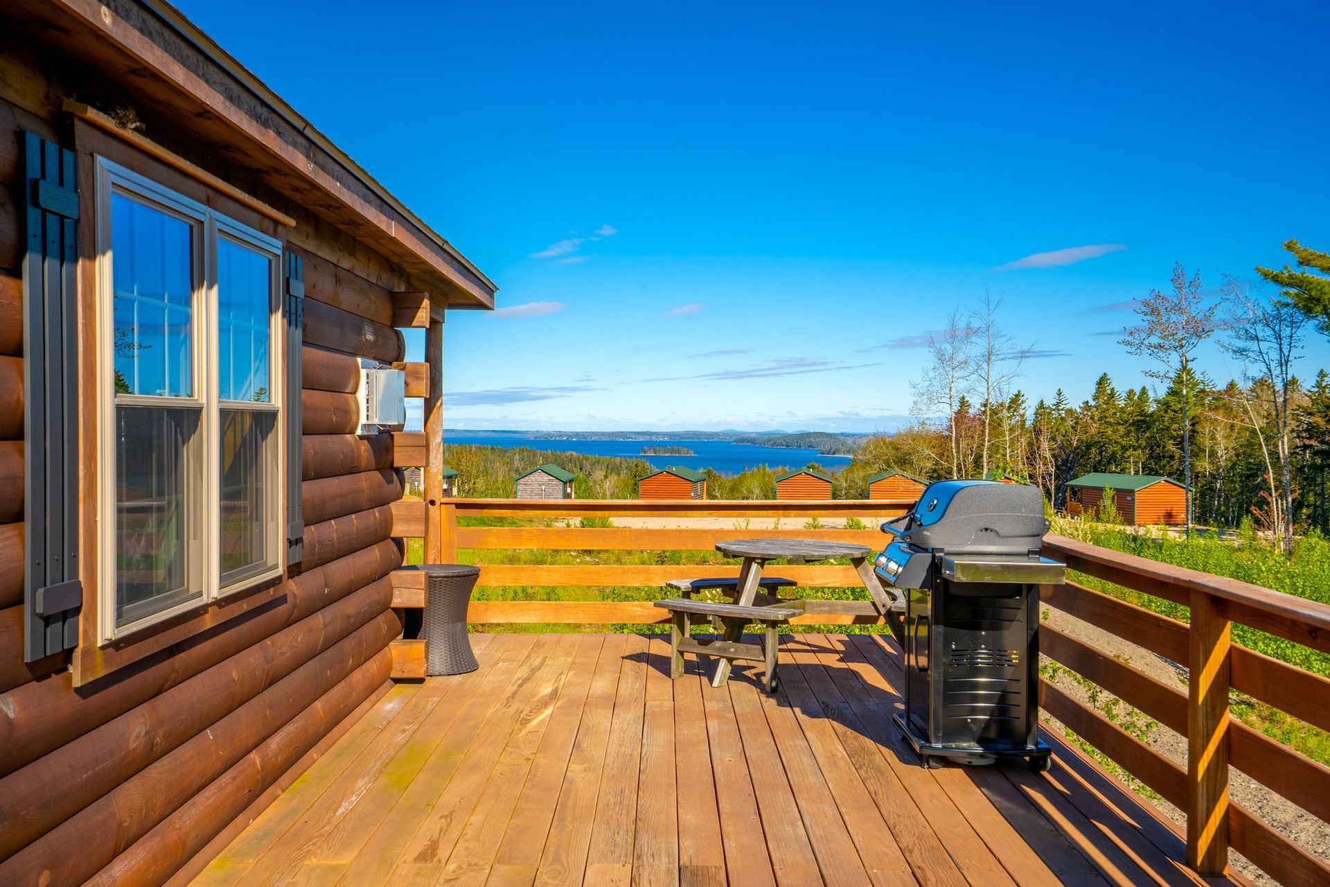 Wooden cabin with a deck overlooking a water view and a picnic table with a grill. Blue sky.