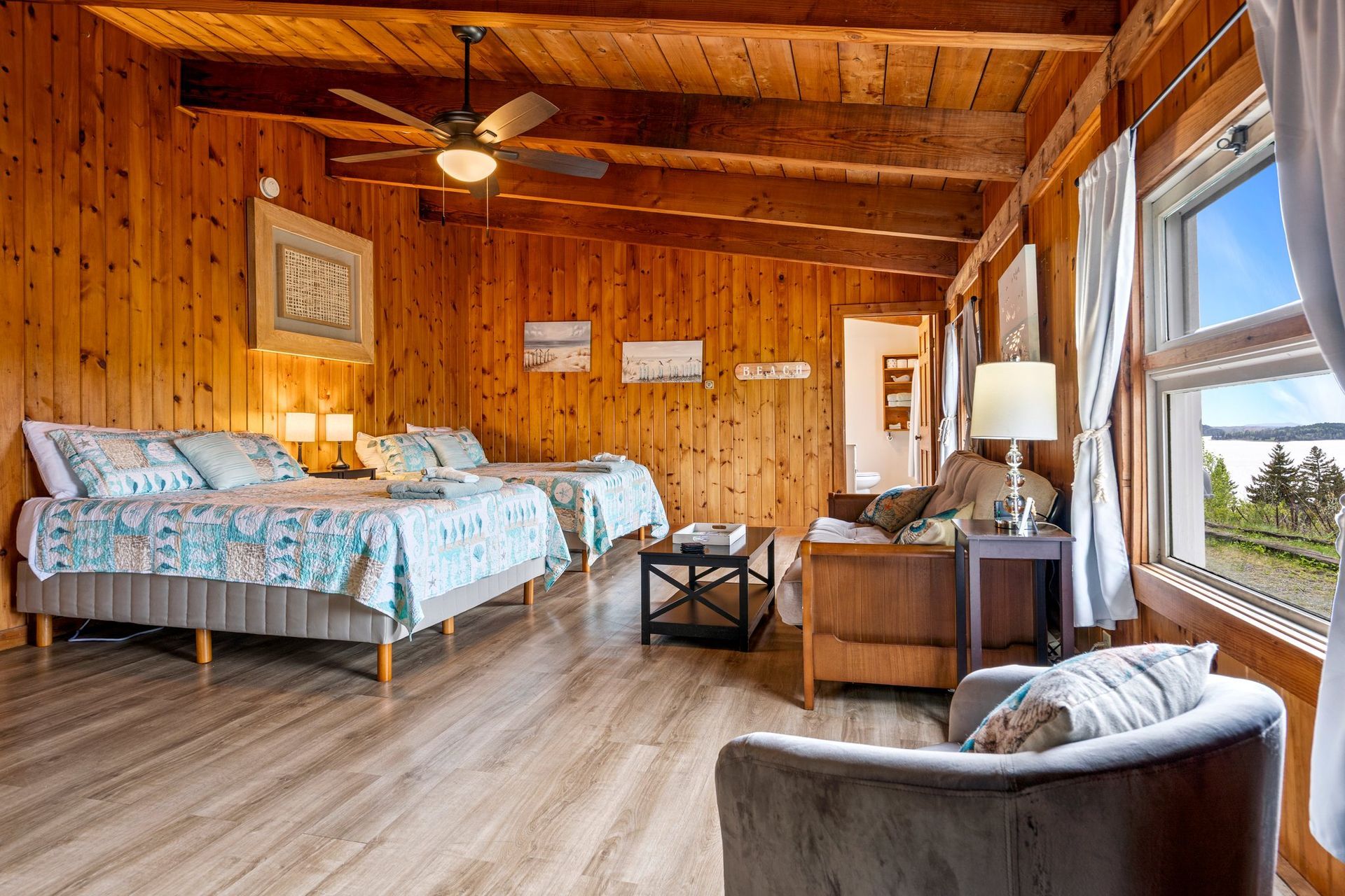 A inn room with wooden paneling, two beds, a seating area, and a window with a view of the outdoors.