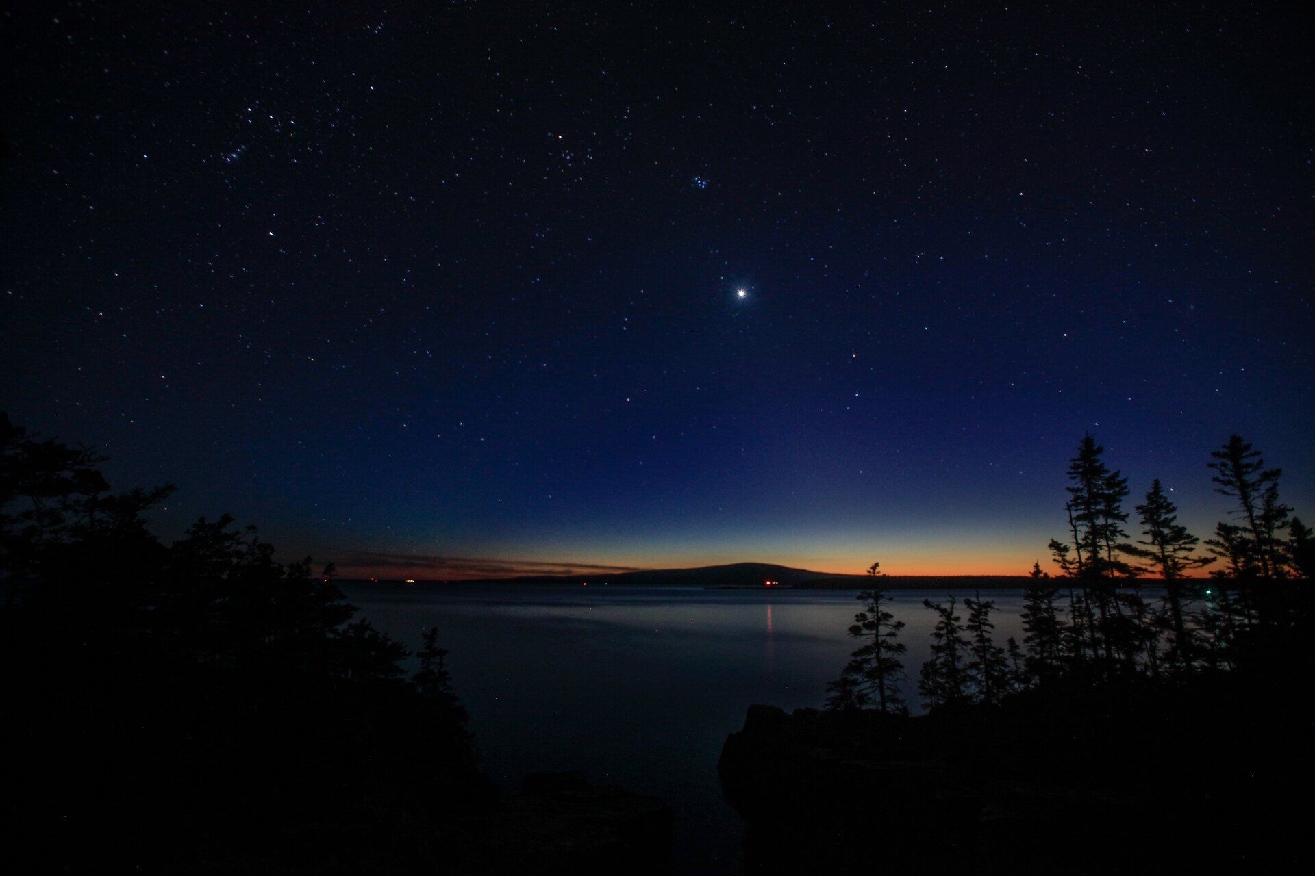 A starry night sky over a lake with trees in the foreground.