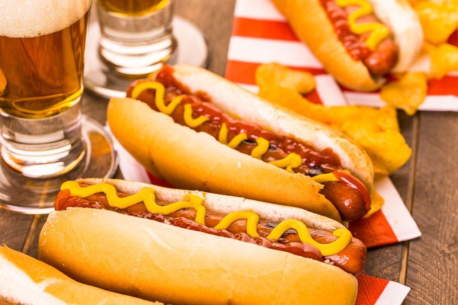 Three hot dogs with mustard and ketchup on a table next to a glass of beer.