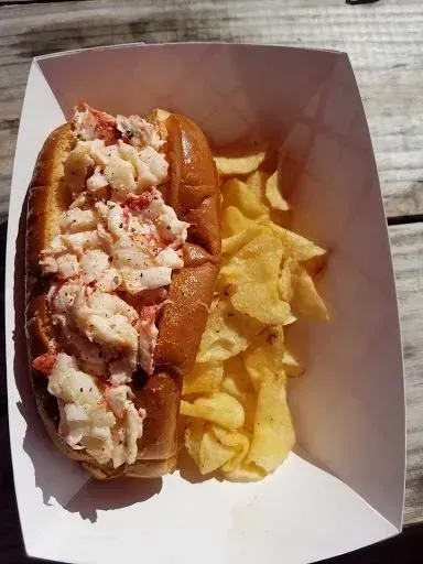 A lobster roll and chips in a paper container on a wooden table.