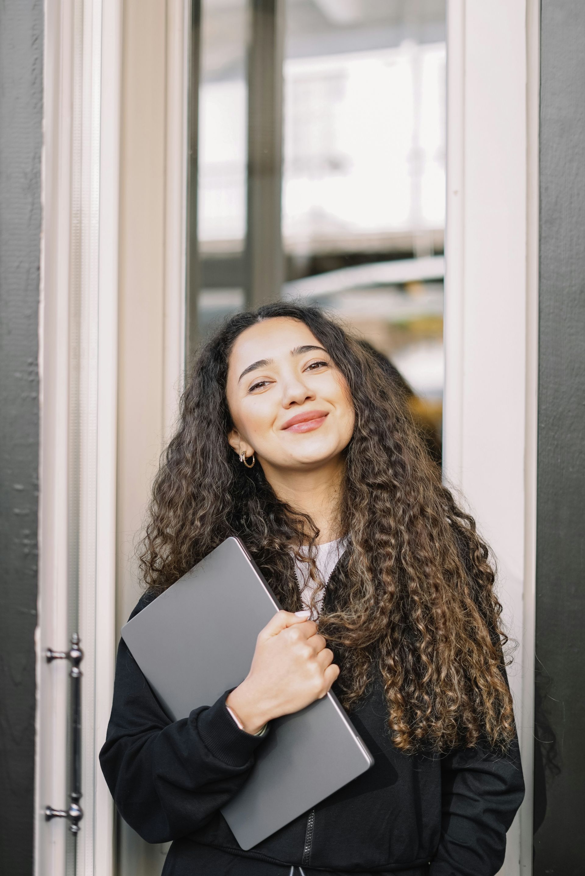 Woman with curly hair smiles while holding a laptop outside a building.