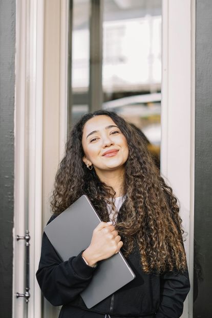 Woman with curly hair smiles while holding a laptop outside a building.