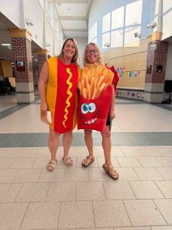 Two women in a hallway wearing a hot dog and french fry costume, smiling at the camera.