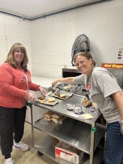 Two women preparing food in a kitchen. One in red, one in gray, smiling. Stainless steel table, buns and toppings visible.