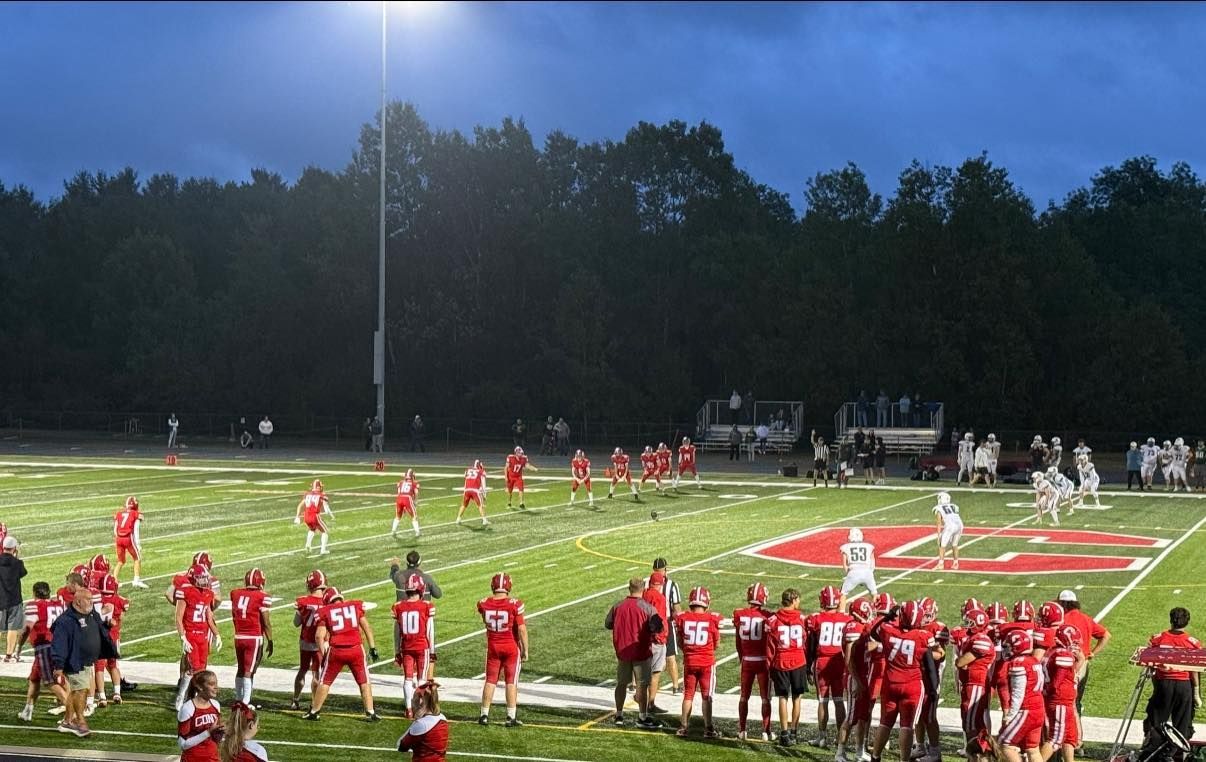 Football game at night under stadium lights; red and white uniforms on the field.