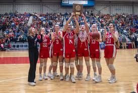 Women's basketball team in red uniforms holding trophy, celebrating victory on court.