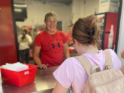 Woman in red shirt smiles at a young person at a food stand.
