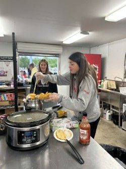 Two young women preparing food in a kitchen; one serves with a ladle, near a slow cooker and a bottle of hot sauce.