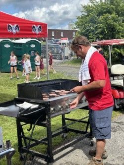 Man grilling burgers outdoors; Red tent, portable toilets, and golf cart in background.