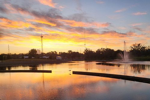 Sunset over a calm pond with pink-orange sky and a fountain spraying in the distance