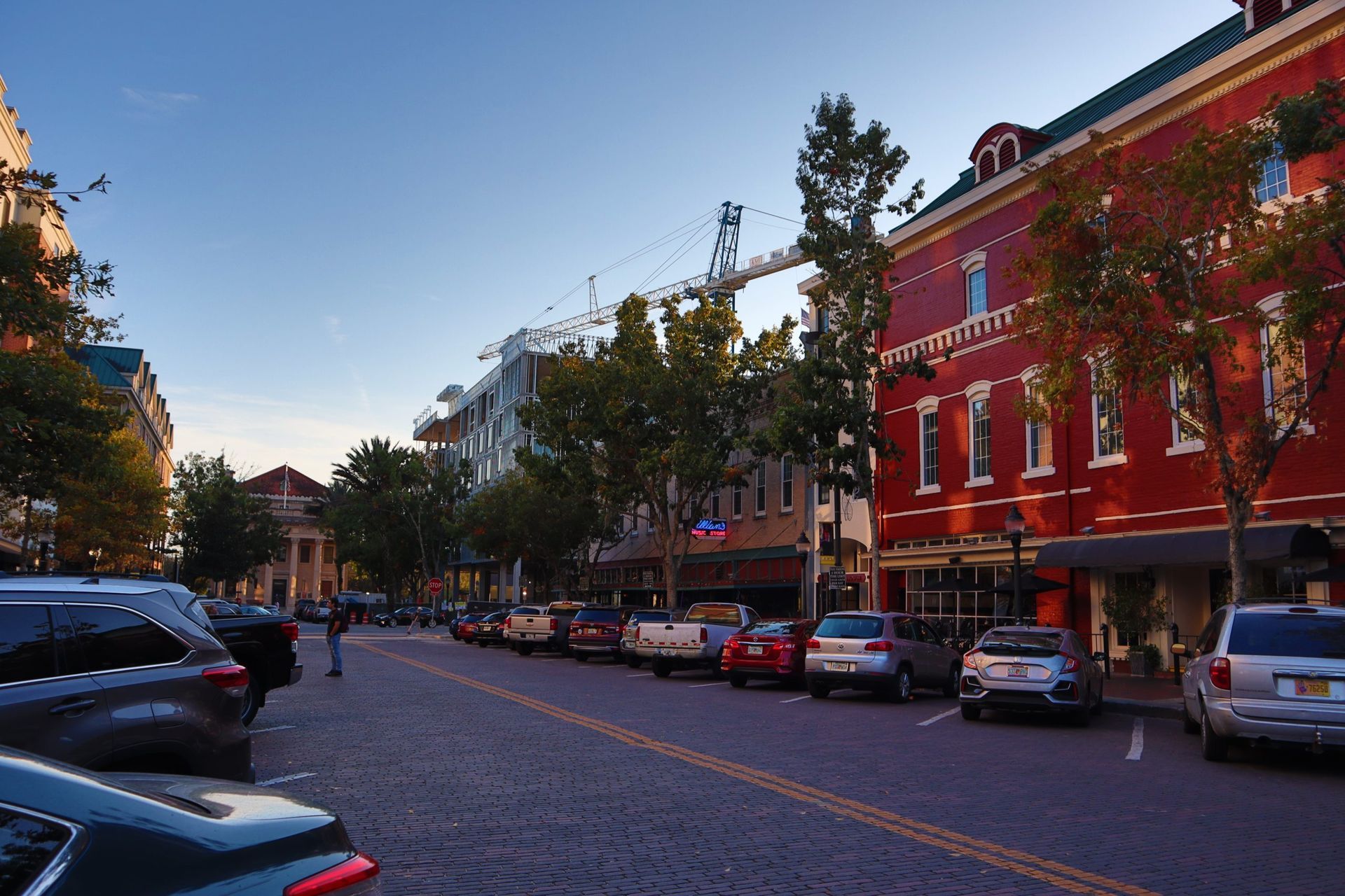 Tree-lined cobblestone street with parked cars and colorful row buildings at dusk
