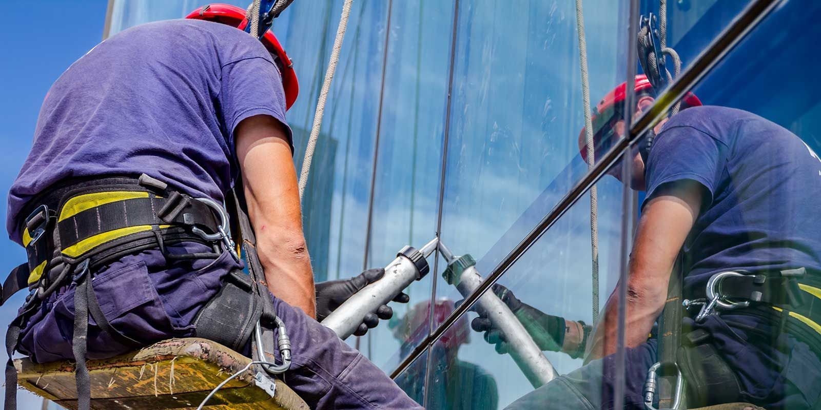 Window washers in harnesses applying sealant to a tall glass building.