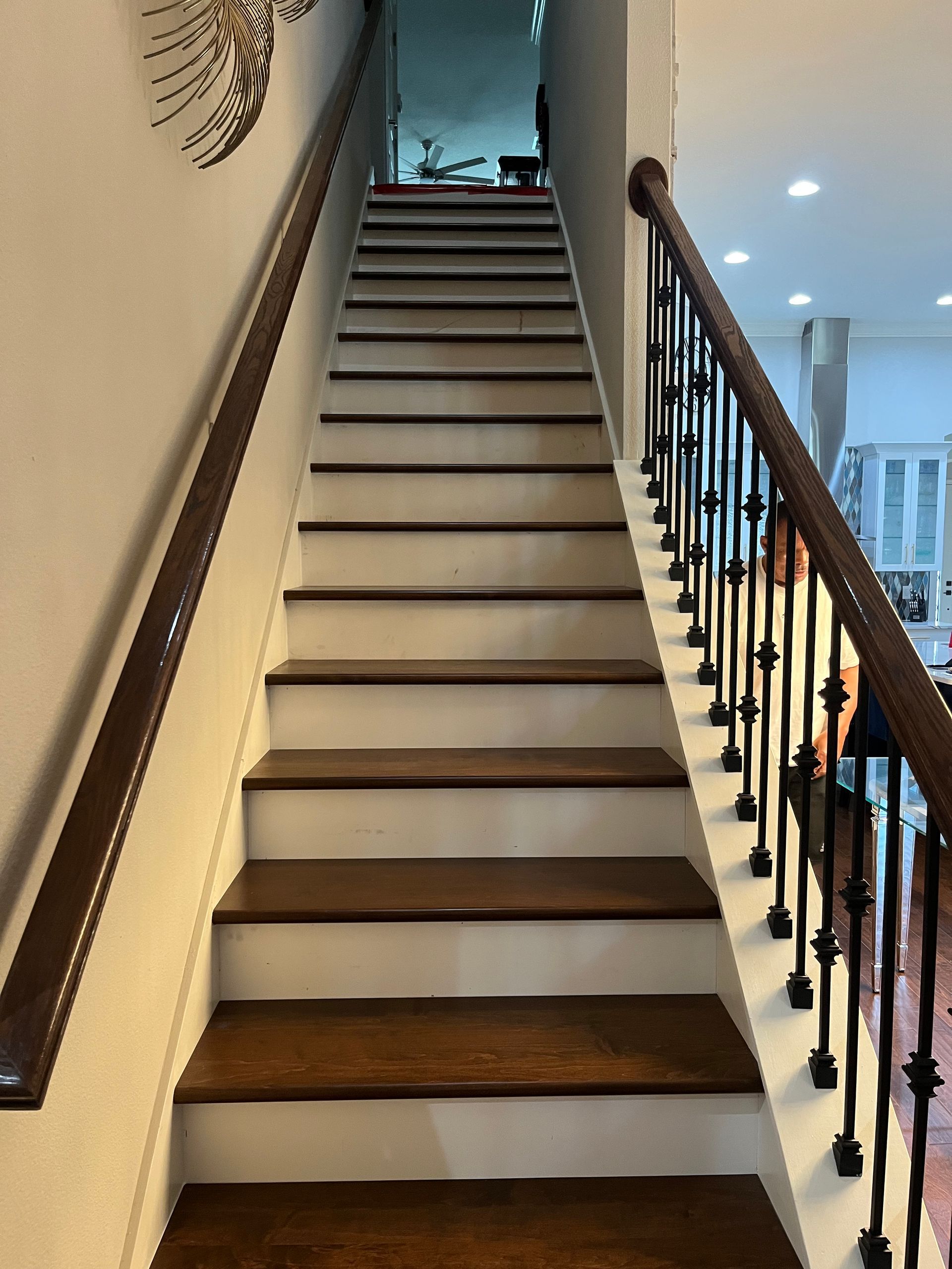 Staircase with brown treads and handrails, black wrought iron railing, white risers, leading upwards to an open doorway.