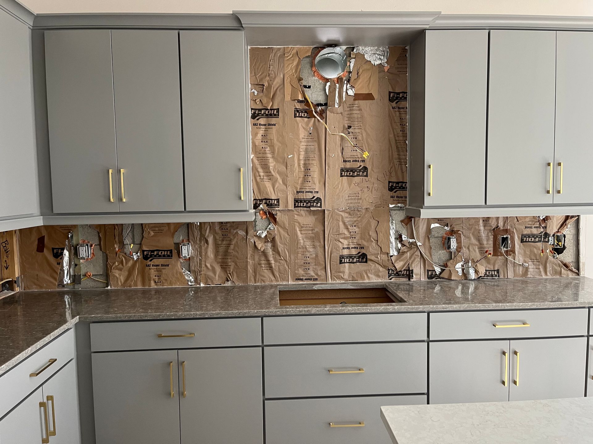 Kitchen cabinets with exposed insulation; renovation in progress. Grey cabinets, tan granite countertop.
