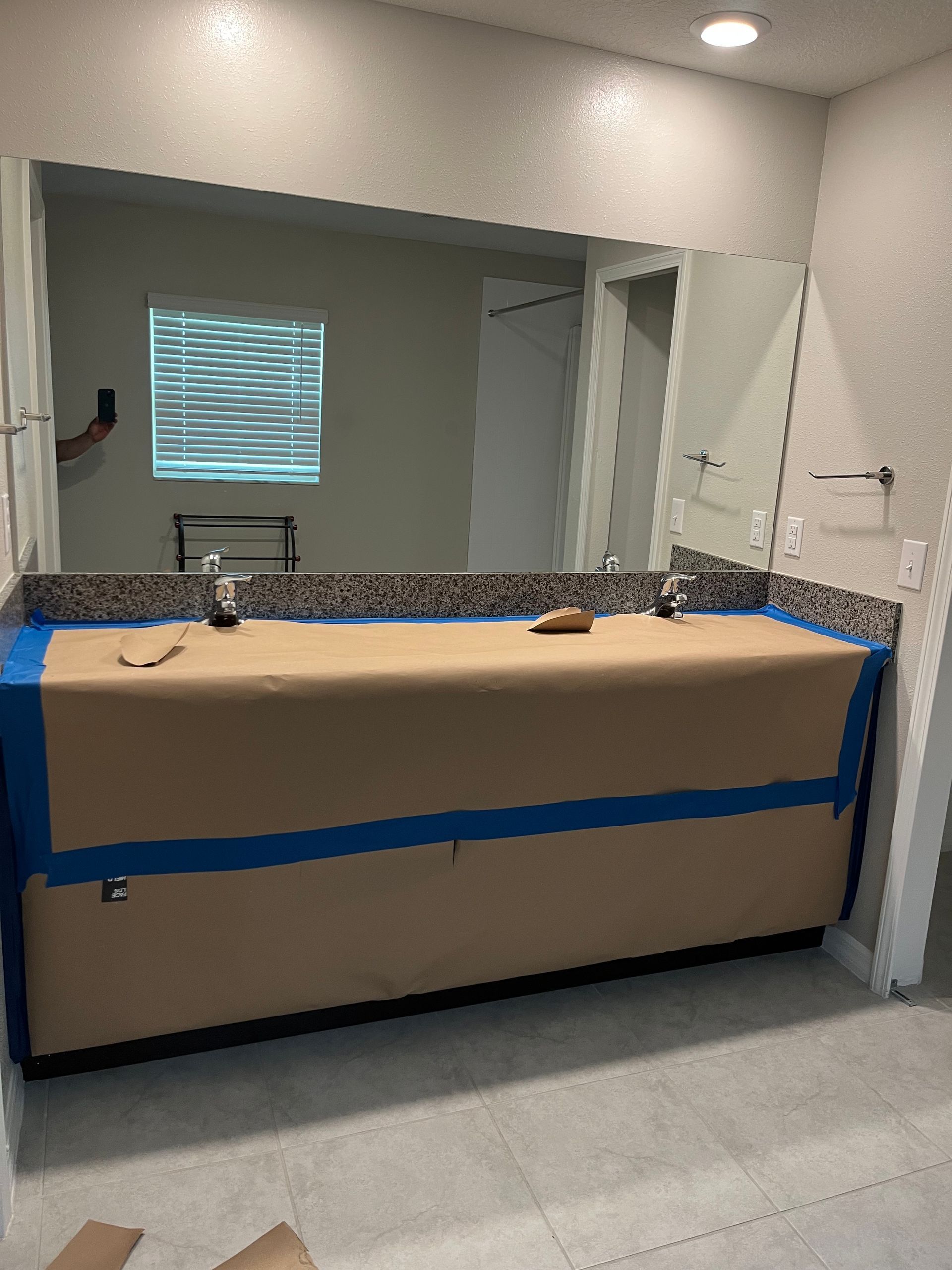 Bathroom vanity covered in brown paper and blue tape, with a large mirror and granite countertop.