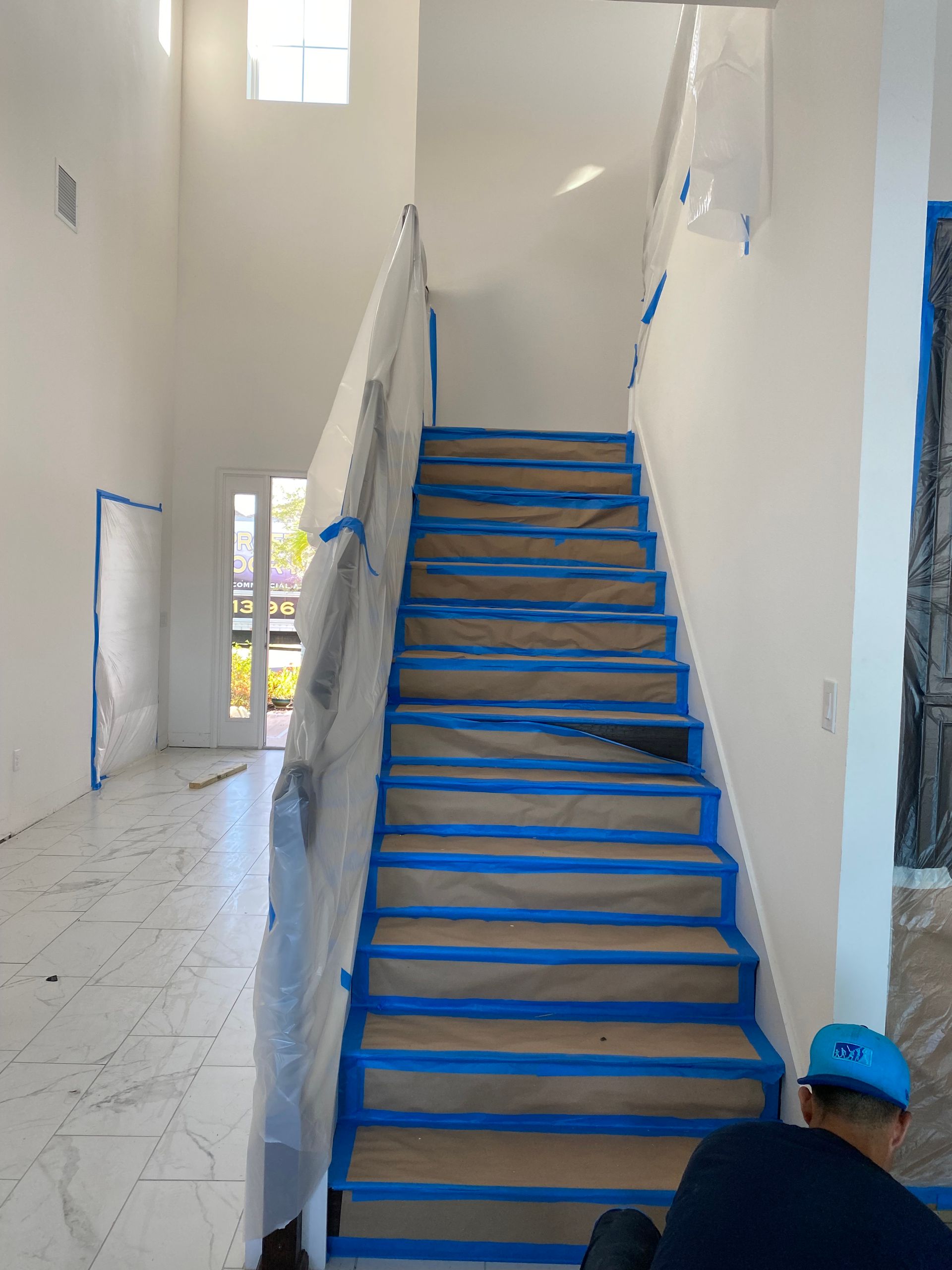 Staircase being prepped for painting. Blue tape covers steps and railings.  Person sits at the bottom. Bright, white walls.