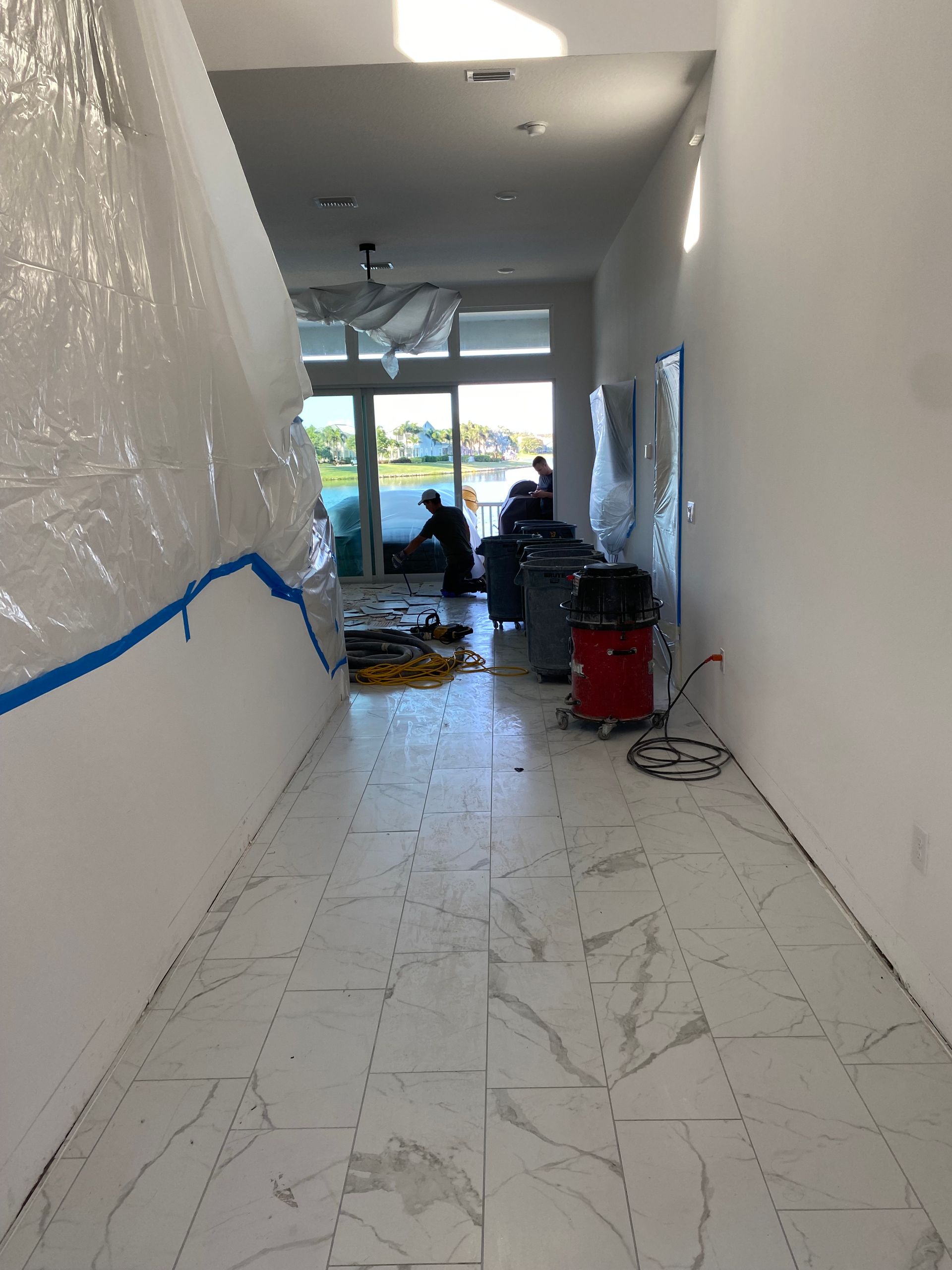 A long hallway under construction with workers, tools, plastic sheeting, and marble tile flooring.