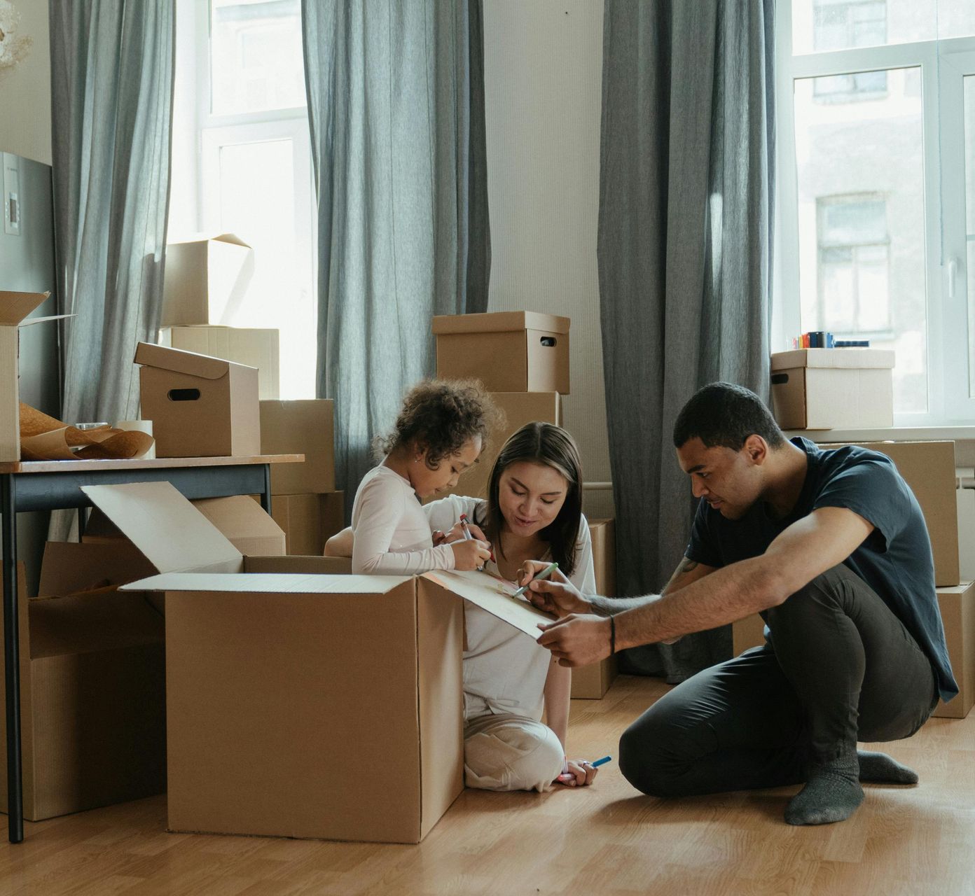 A family is sitting on the floor in a room filled with cardboard boxes.