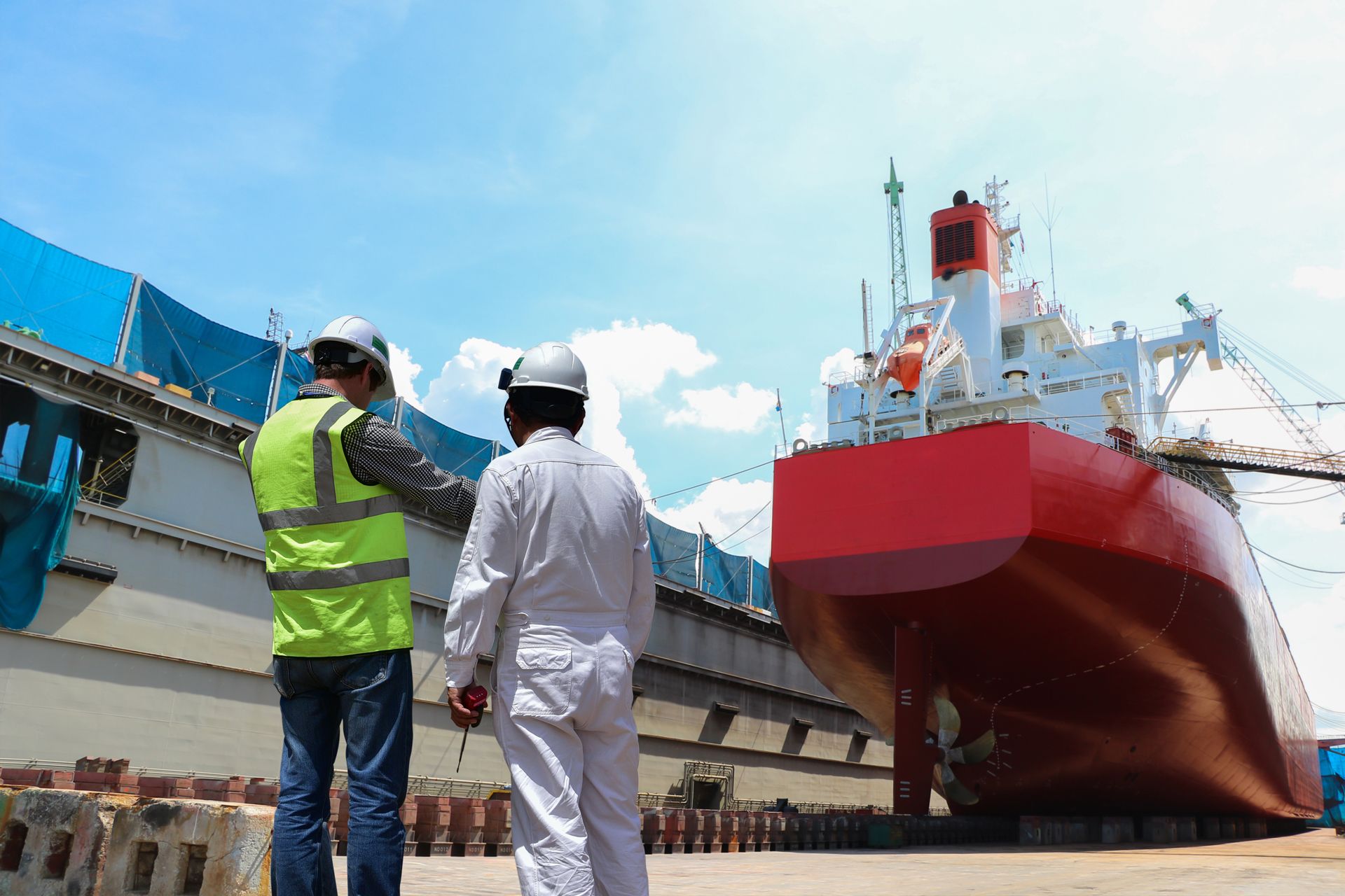 Two shipyard workers inspect a red cargo ship in drydock