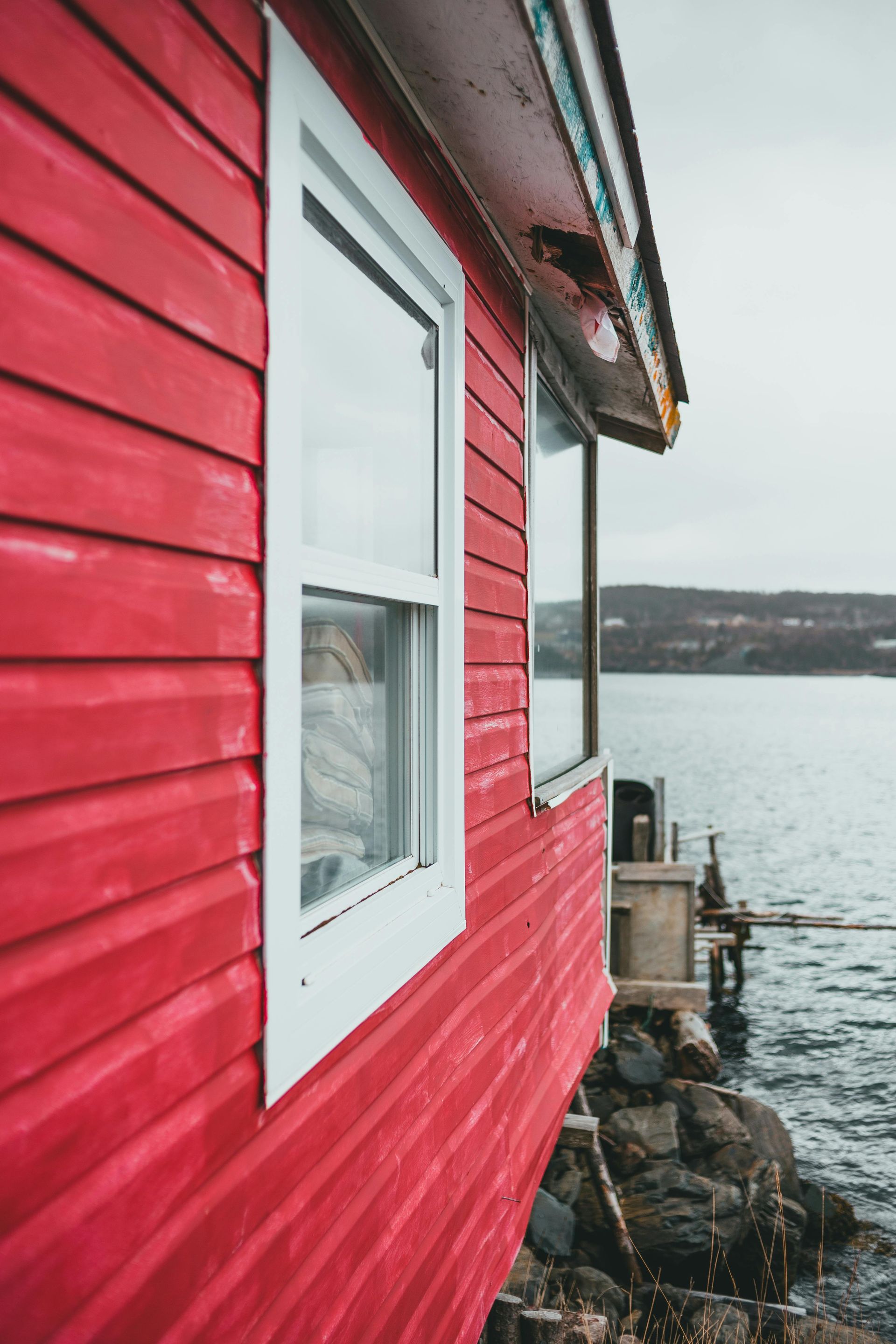 Red wooden house with white-trimmed window beside a calm waterfront under an overcast sky