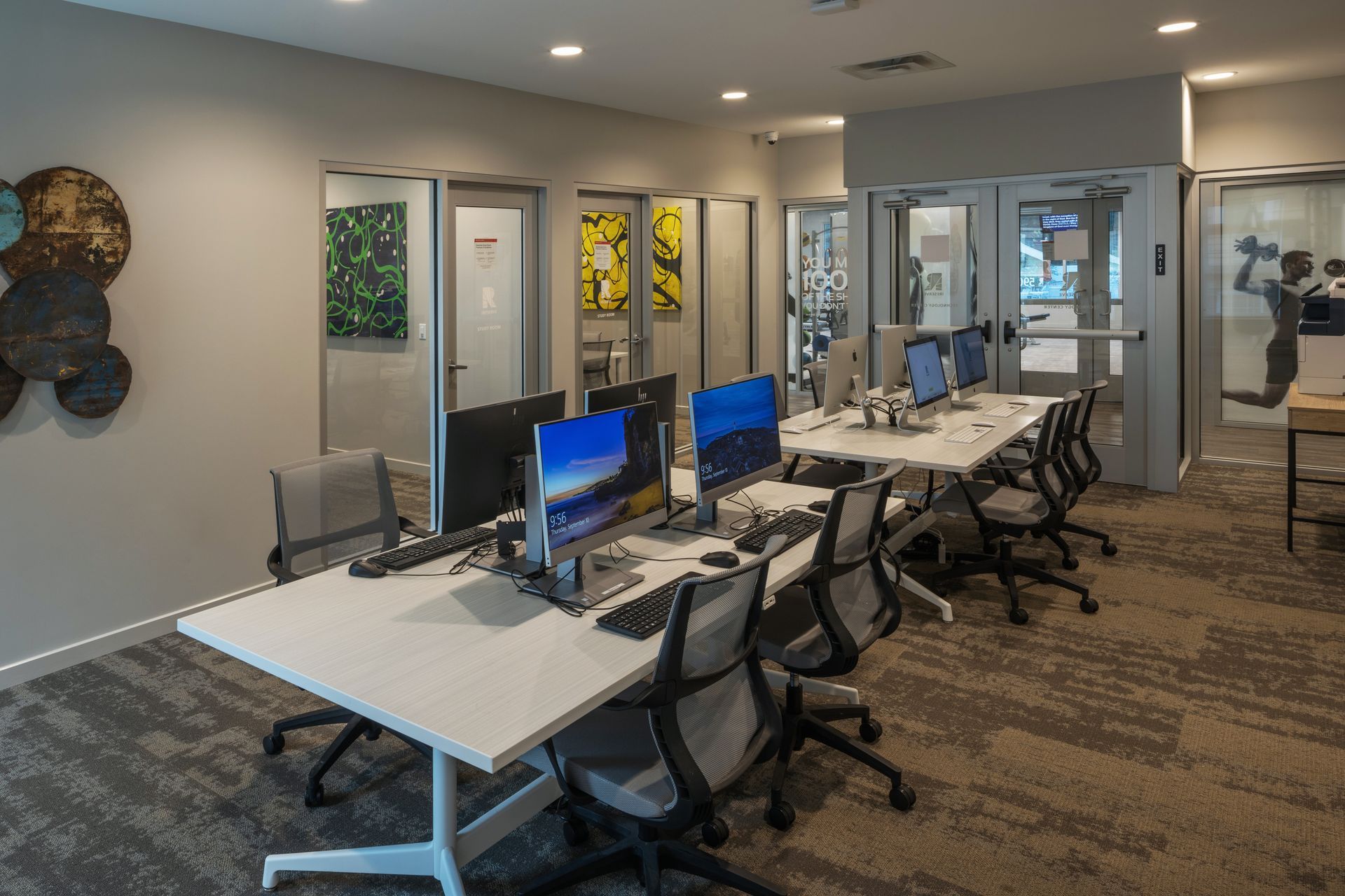 Computer lab with long white desks, monitors, and rolling chairs. Doors with artwork line the back wall.