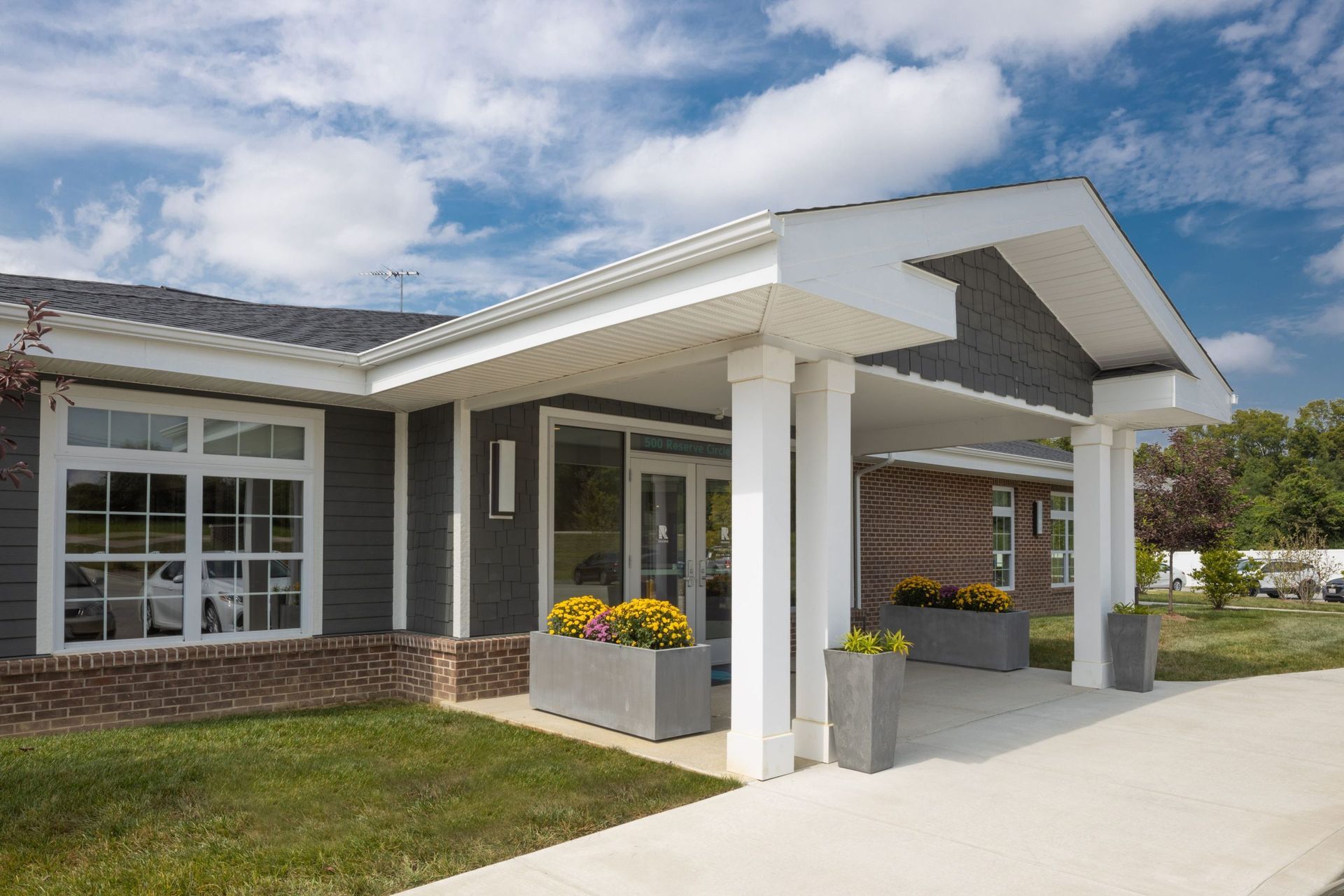 Exterior of a building with a covered entrance. Gray siding and brick with flower planters and columns.
