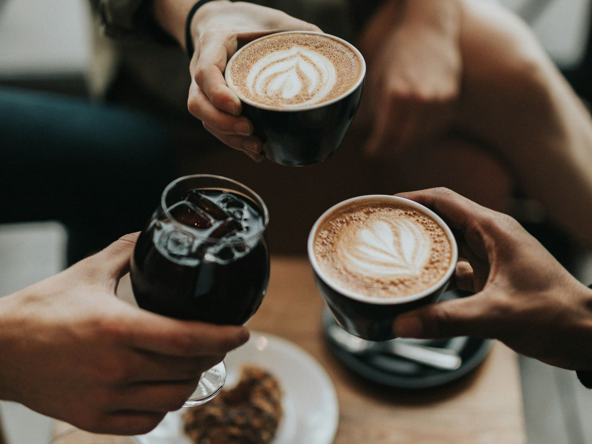Three people holding coffee and a glass of iced coffee, clinking together.