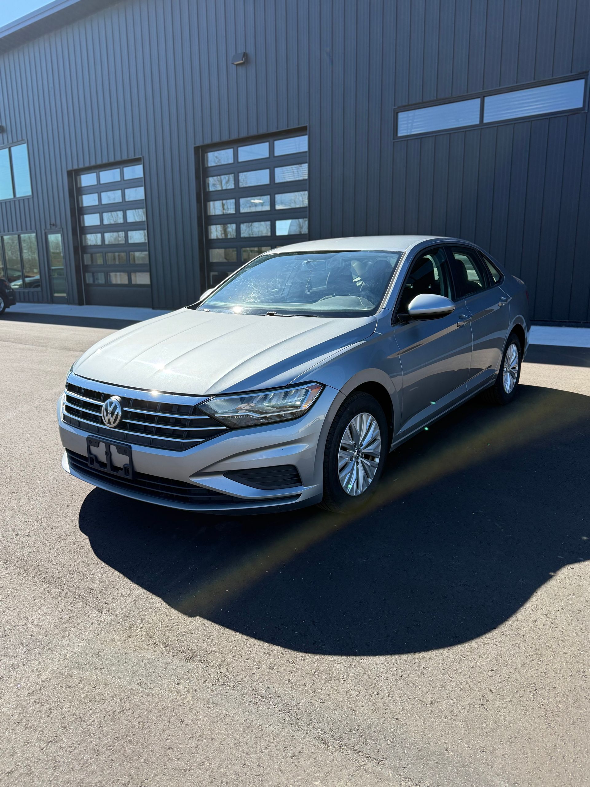 A silver Volkswagen Jetta parked in front of a dark gray building with glass pane garage doors on a sunny day.