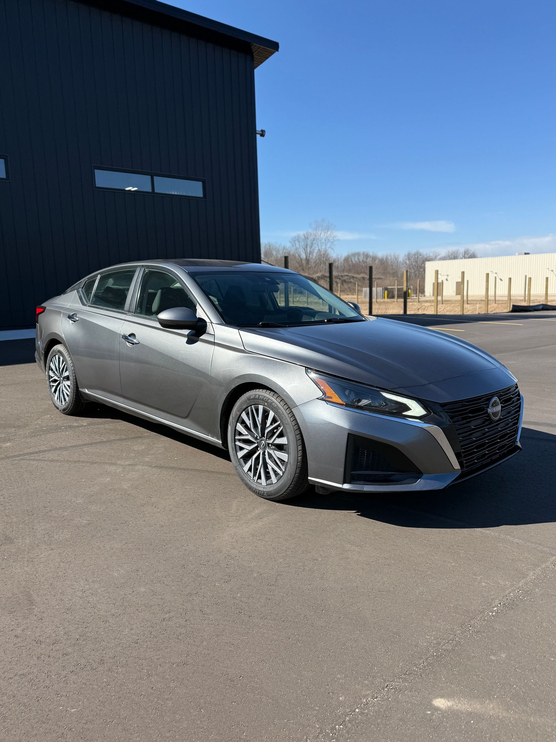 A gray Nissan Altima parked on an asphalt lot in front of a dark, industrial-style building under a clear blue sky.