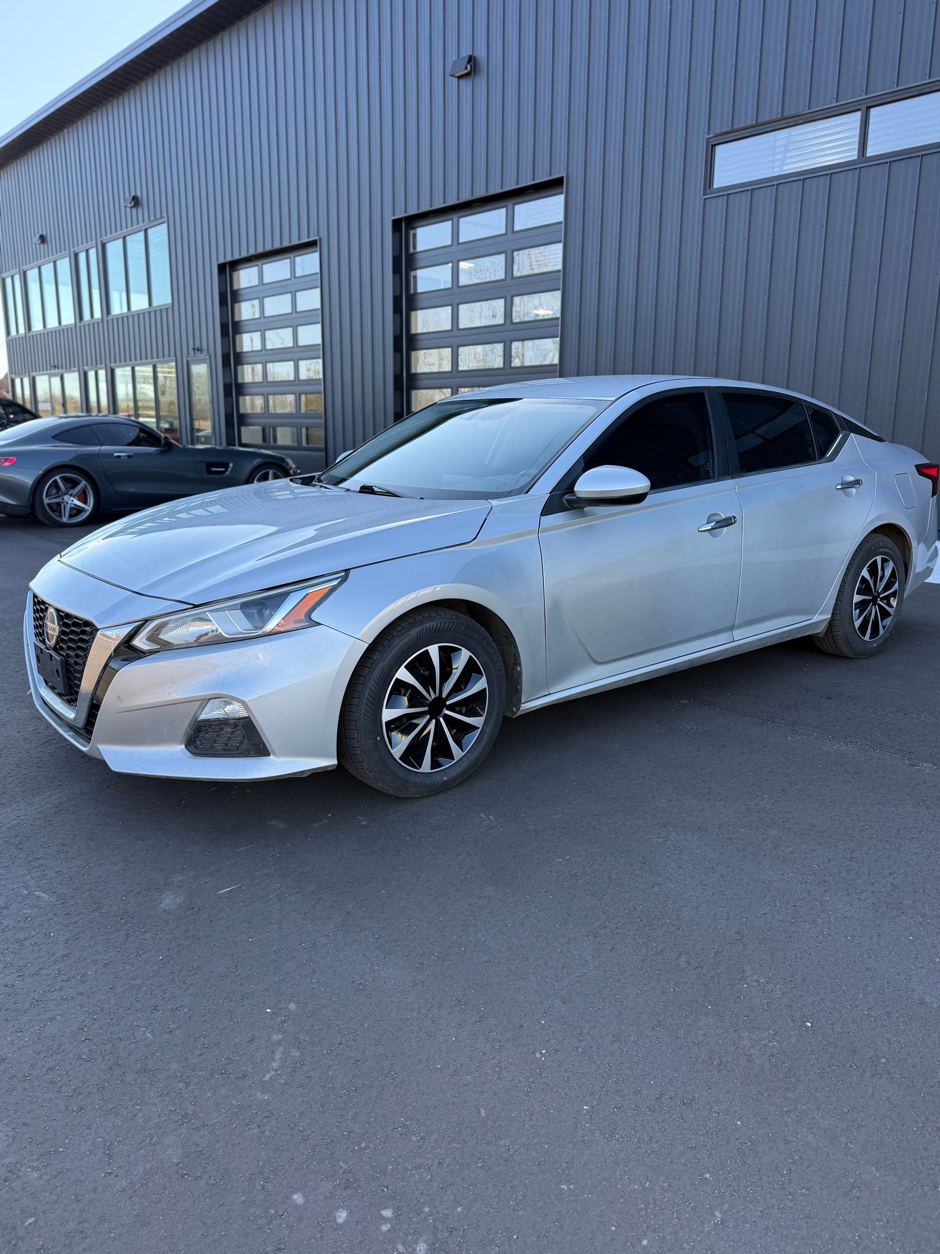 A silver Nissan Altima parked in front of a dark gray modern industrial building.