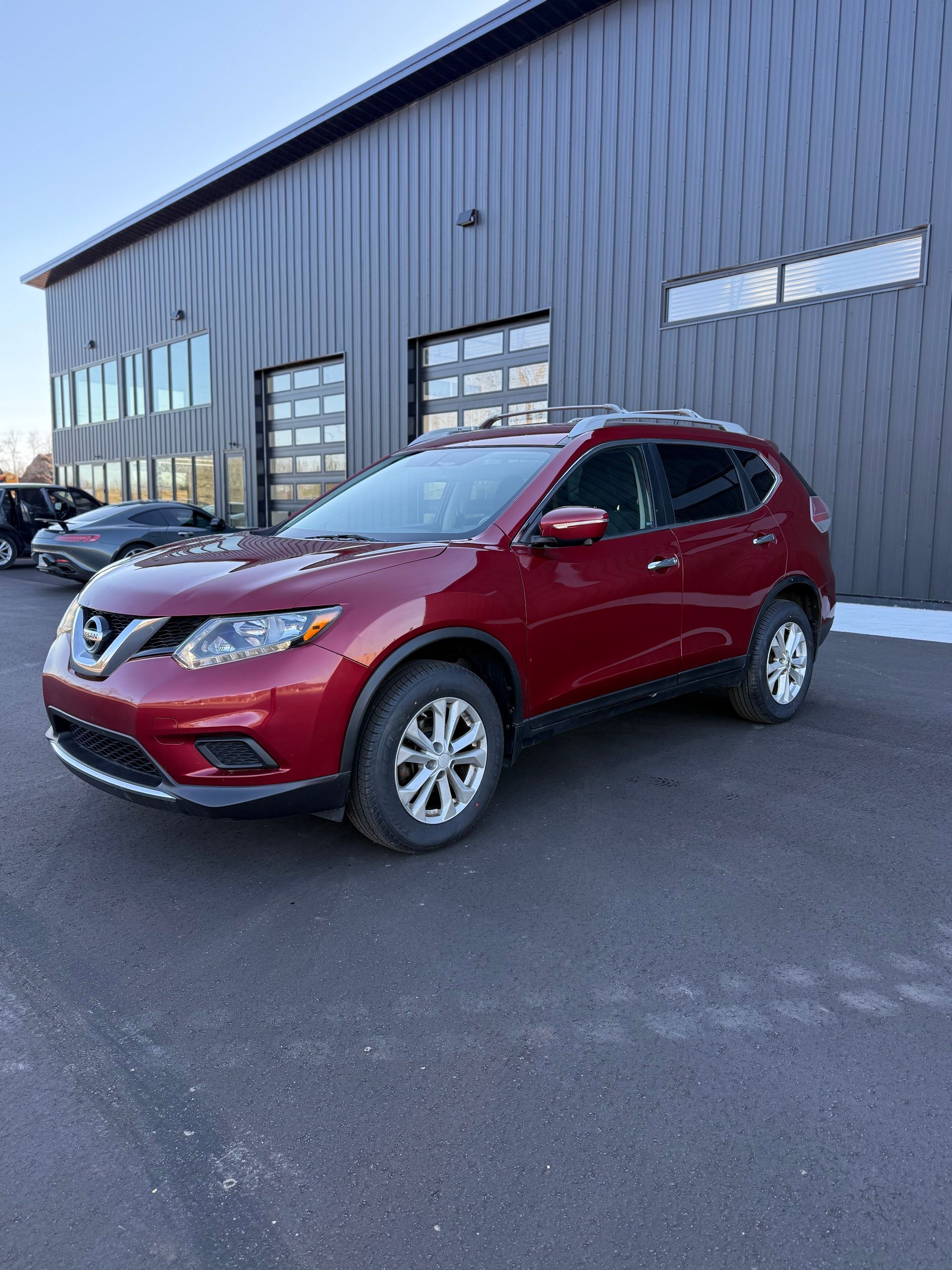 A red Nissan Rogue parked on asphalt in front of a modern industrial building with black metal siding.