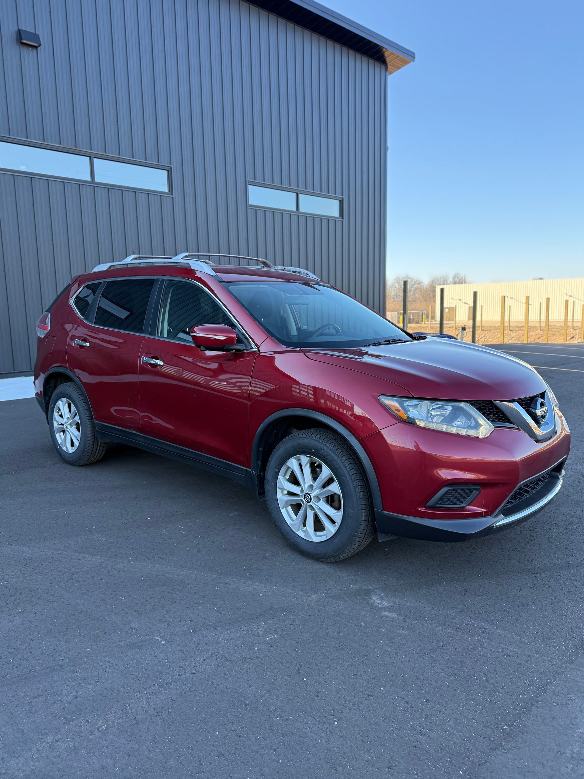 A red Nissan Rogue parked on asphalt in front of a modern industrial building with black metal siding.