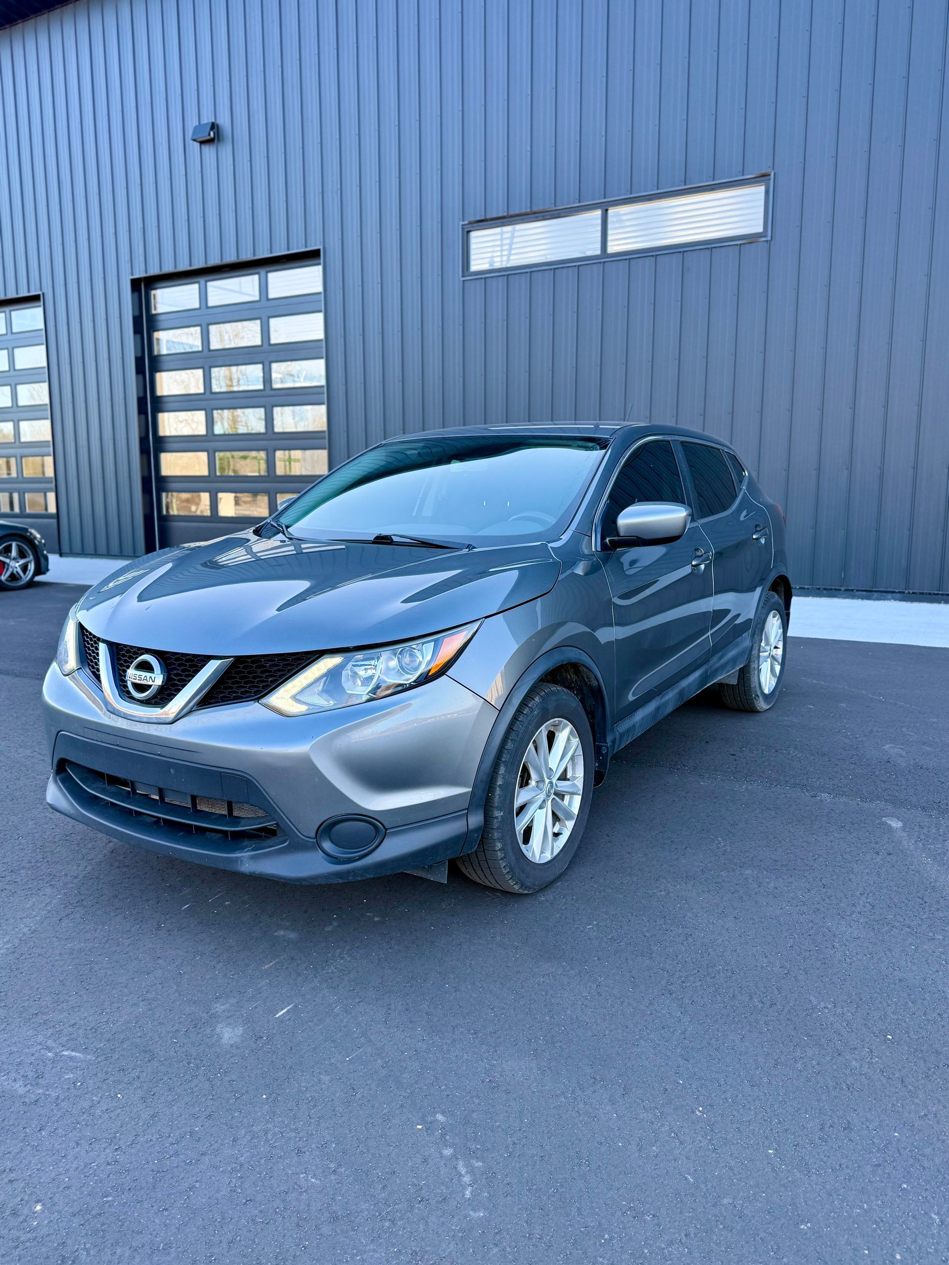 A gray Nissan Rogue parked on an asphalt lot in front of a dark-paneled industrial building.